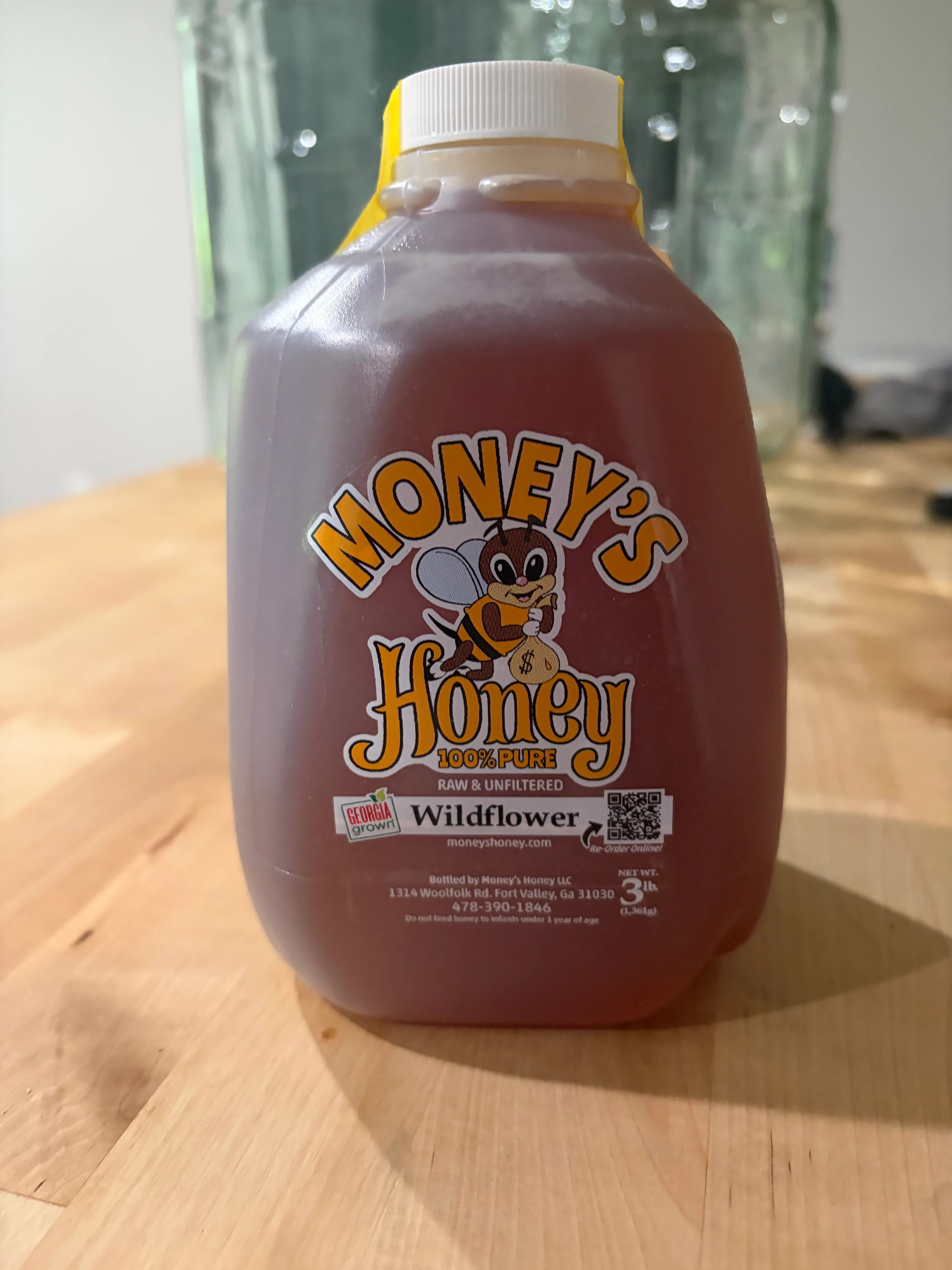 A jug of Money's Honey Wildflower honey on a wooden counter, glass carboys behind it.
