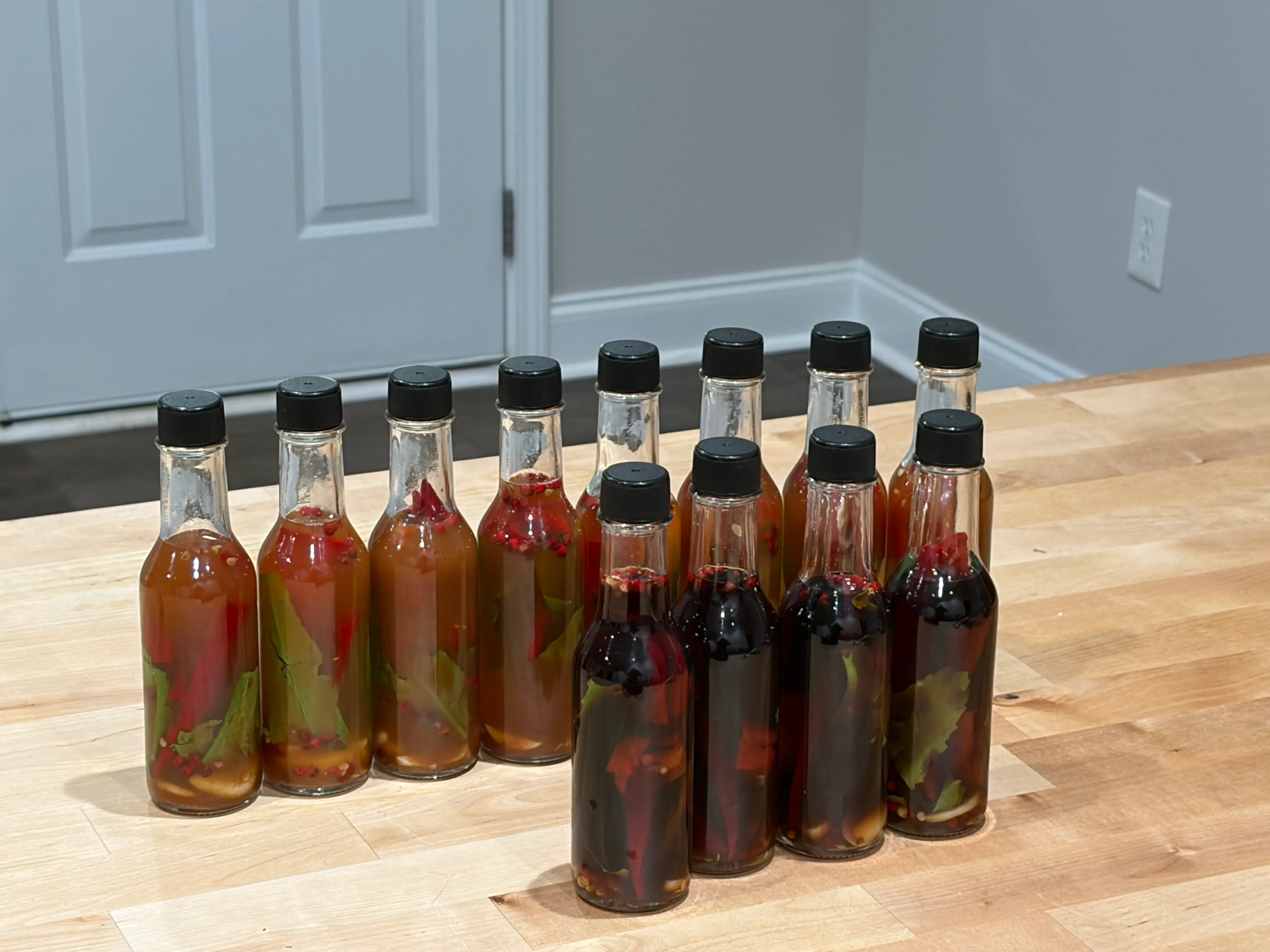 A dozen small glass bottles with black caps — some filled with amber vinegar, some deep red — each packed with whole red chilis, garlic, and green culantro leaves.