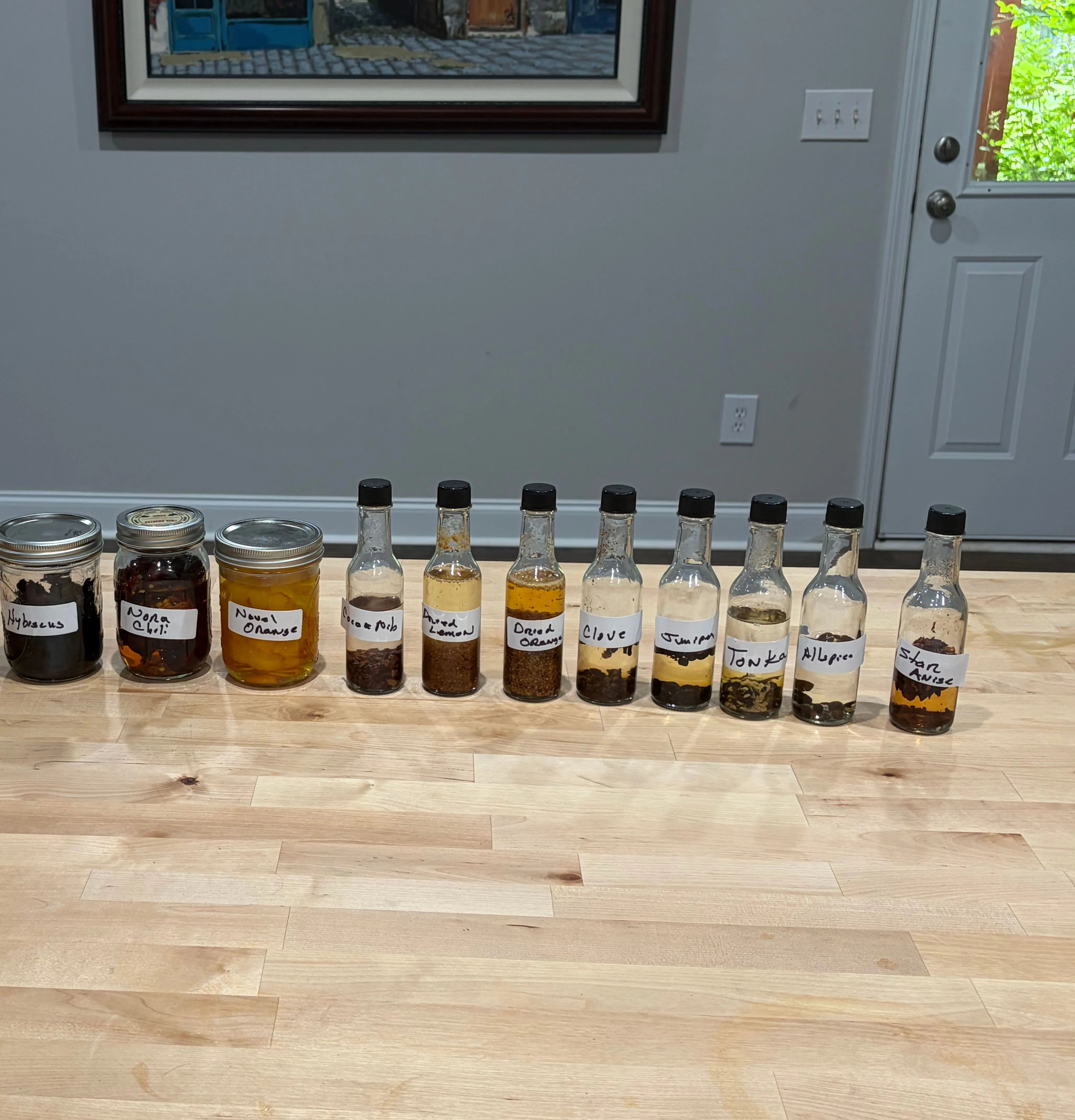 A row of labeled jars and small bottles — Hibiscus, Nora Chili, Navel Orange, Cocoa Nib, Clove, Juniper, Tonka, Allspice, Star Anise and more — each steeping in vodka.